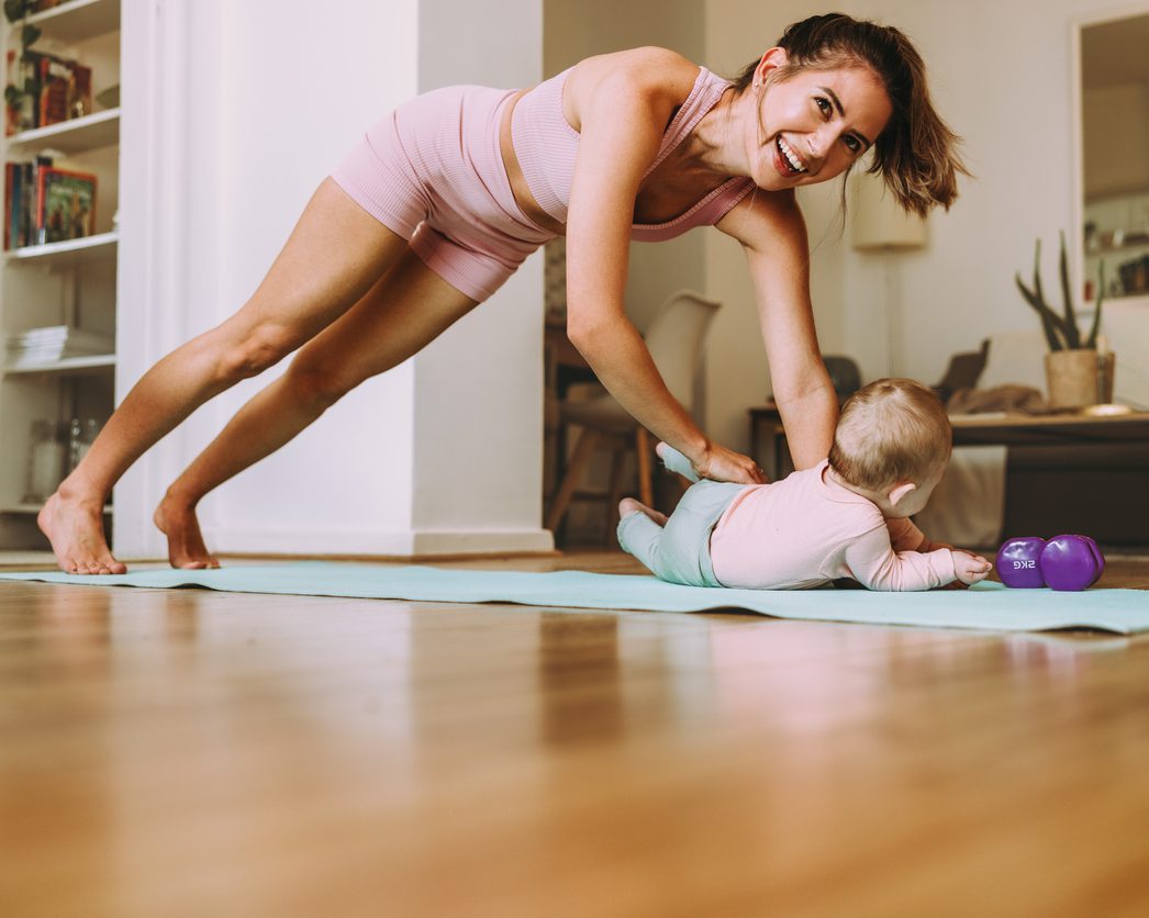 Happy young mom working out with her baby at home. Cheerful mom smiling while doing push up exercises on an exercise mat. New mom bonding with her baby during her post-natal fitness routine.